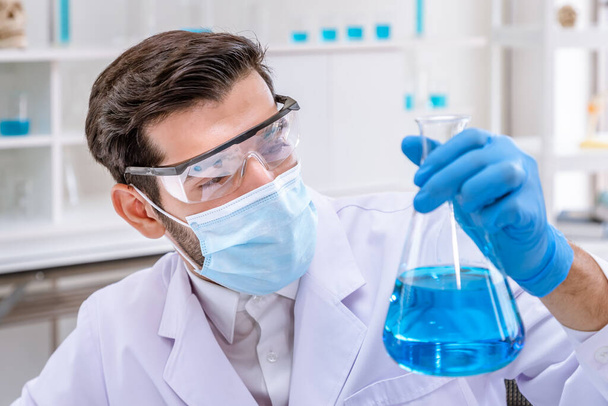 Άραβας αρσενικός ερευνητής που κρατάει και εξετάζει τη φιάλη Erlenmeyer γεμάτη με μπλε υγρή χημική ουσία στο εργαστήριο - Φωτογραφία, εικόνα