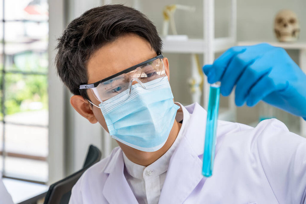 Asyalı erkek araştırmacı bilim adamı laboratuvarda mavi sıvı kimyasallarla dolu test tüpünü tutuyor ve inceliyor. - Fotoğraf, Görsel