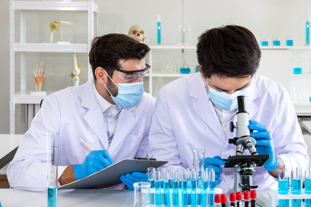 Due ricercatori maschi asiatici e arabi che lavorano in laboratorio, conducendo studi sulla sostanza a rischio biologico con attrezzature scientifiche e microscopio - Foto, immagini