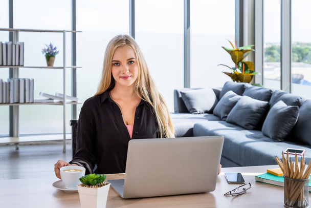 Portret van een jonge blanke vrouw met blond haar freelancer thuis, zittend aan een bureau in de woonkamer, kijkend naar de camera - Foto, afbeelding