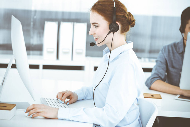 Young woman using headset and computer while talking with customers online. Group of Call center operators at work. Business concept - Foto, afbeelding
