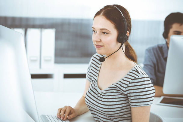 Casual dressed young woman using headset and computer while talking with customers online. Group of operators at work. Call center, business concept - Foto, Imagen