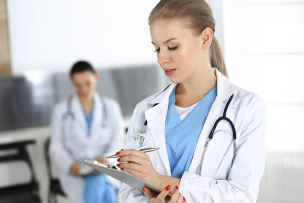 Woman - doctor is using clipboard for filling up medication history record in clinic with colleague at the background - Photo, image