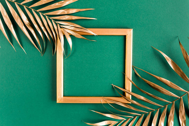 Gold tropical palm leaves and photo frame on green background. Flat lay, top view minimal concept. - Фото, изображение