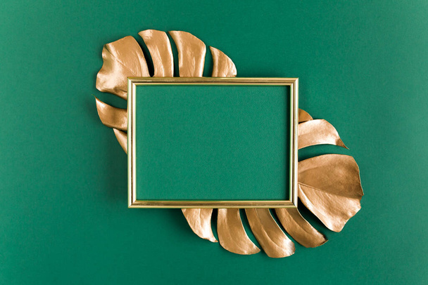 Gold tropical palm leaves Monstera and photo frame on green background. Flat lay, top view minimal concept. - Fotó, kép