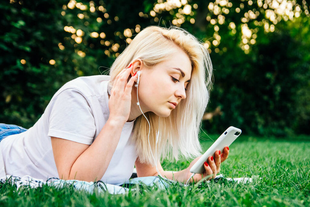 Beautiful blonde woman in headphones and with a smartphone in his hands listening to music lying on grass outdoors in park. Place for text or advertising. - Valokuva, kuva