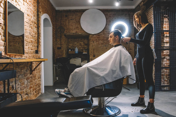 Όμορφη κομμώτρια κόβει τα μαλλιά του πελάτη με ψαλίδι σε κουρείο. Έννοια της διαφήμισης και της κομμωτικής. - Φωτογραφία, εικόνα