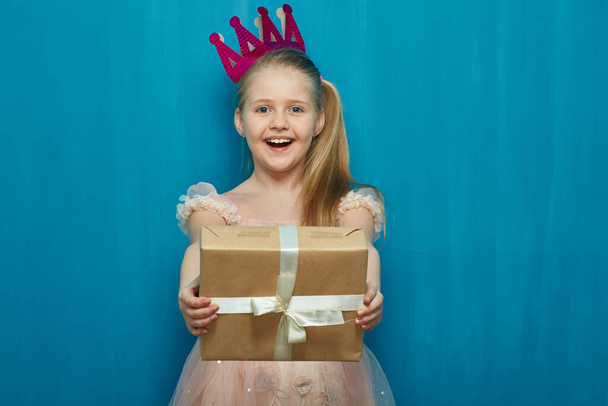 Ευτυχισμένο κορίτσι παιδί φορώντας ροζ φόρεμα και το στέμμα κρατώντας κουτί δώρου. - Φωτογραφία, εικόνα