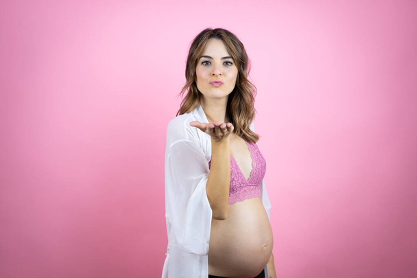 Young beautiful brunette woman pregnant expecting baby over isolated pink background looking at the camera blowing a kiss with hand on air being lovely and sexy. Love expression. - Fotó, kép