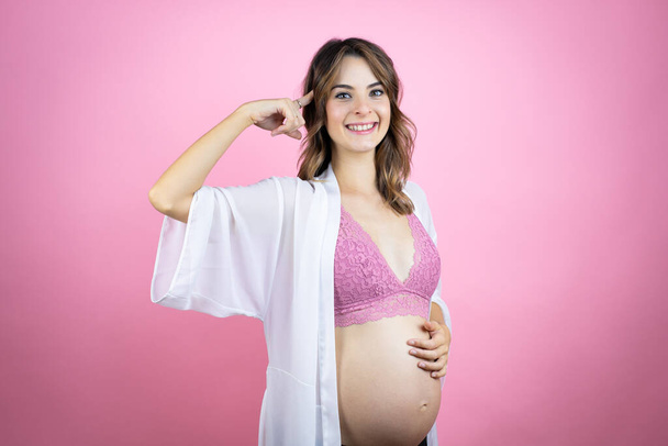 Νεαρή όμορφη μελαχρινή γυναίκα έγκυος περιμένει μωρό πάνω από απομονωμένο ροζ φόντο χαμογελώντας και σκέφτεται με τα δάχτυλά της στο κεφάλι της ότι έχει μια ιδέα. - Φωτογραφία, εικόνα