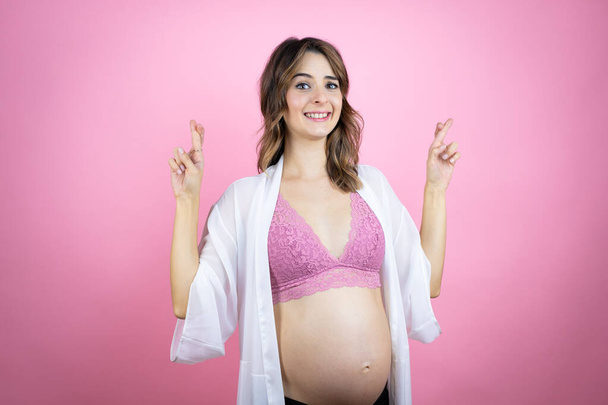 Nuori kaunis brunette nainen raskaana odottaa vauvan yli eristetty vaaleanpunainen tausta ele sormi ristissä hymyillen toivoa ja etsivät puolella - Valokuva, kuva