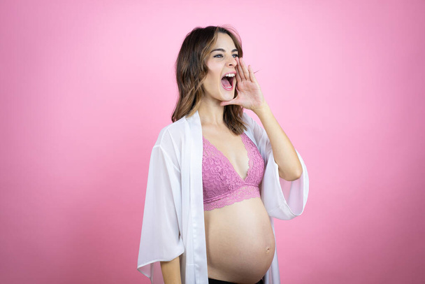 Jovem bela mulher morena grávida esperando bebê sobre fundo rosa isolado gritando e gritando alto para o lado com a mão na boca - Foto, Imagem