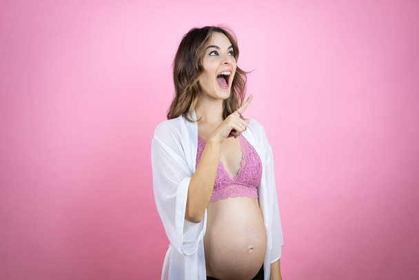 Giovane bella donna bruna incinta in attesa di bambino su sfondo rosa isolato sorridente e indicante con mano e dito di lato - Foto, immagini