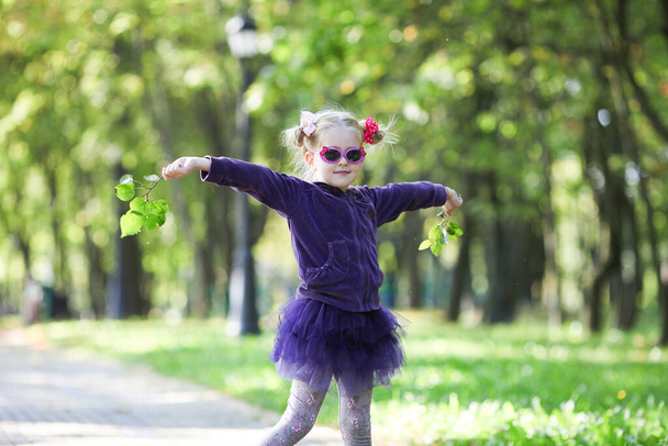 Γλυκό κοριτσάκι με κουλούρια που φοράει υπέροχα γυαλιά ηλίου και φούστα tutu παίζοντας με πράσινα κλαδιά δέντρων στο πάρκο. Ευτυχισμένη παιδική ηλικία, αγάπη άνοιξη έννοια.  - Φωτογραφία, εικόνα
