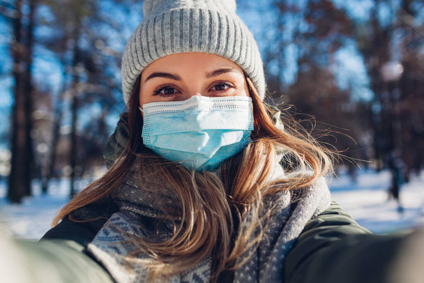 雪の冬の公園で護身用フェイスマスクをした女性が自撮り。コロナウイルスcovid-19パンデミック屋外安全対策. - 写真・画像