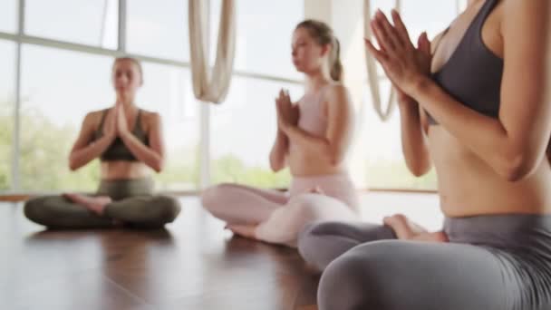 Valkoihoiset tytöt, jotka istuvat lattialla lootuksessa, aiheuttavat meditointia silmät kiinni tyhjässä joogastudiossa, jossa on panoraamaikkunat. - Materiaali, video