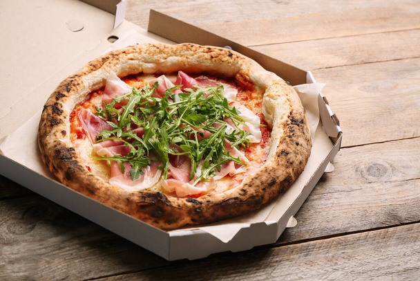 Lekkere pizza met vlees en arugula in kartonnen doos op houten tafel - Foto, afbeelding