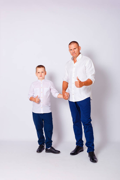 father holds his nine-year-old son's hand. man and boy in white shirts on a white background - Photo, image