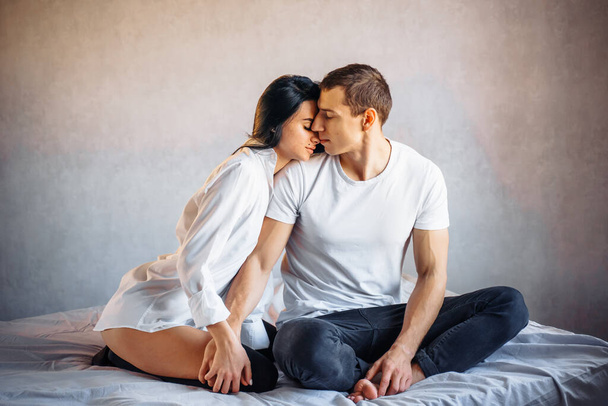 Μια παθιασμένη γυναίκα αγκαλιάζει έναν άντρα τρυφερά και τον φιλάει ρομαντικά. Νεαρός ευγενικός εραστής, κάθεται στο κρεβάτι, απολαμβάνοντας το άγγιγμα της αισθησιακής σέξι κυρίας, γκρίνια με την επιθυμία κατά τη διάρκεια του σεξ. - Φωτογραφία, εικόνα