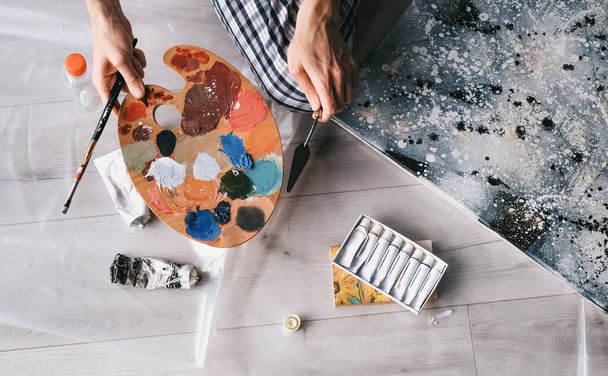Der junge Mann mischt Ölfarben auf einer Holzpalette, um zu Hause ein Bild der Selbstisolierung zu malen. Kunsttherapie. Freizeit zu Hause. Kunstworkshop - Foto, Bild