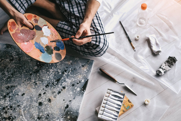 Молодой человек смешивает масляные краски на деревянной палитре, чтобы нарисовать картину дома при самоизоляции. арт-терапия. домашний отдых. арт мастерская - Фото, изображение