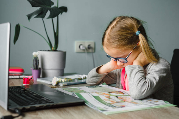 Kleines Mädchen in Augengase schaut sich ihr Klassenbuch an, während sie zu Hause Online-Unterricht hat - Foto, Bild
