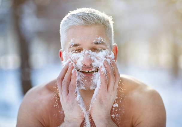 Pozitivní zralý chlap si myje obličej sněhem, stojí s holým trupem v zimním lese, vyvíjí odolnost vůči chladu - Fotografie, Obrázek