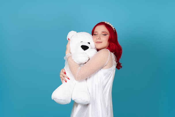 a beautiful young woman in a white dress with red hair with her eyes closed gently presses her cheek to the muzzle of a white teddy bear, isolated on a blue background - Foto, Bild