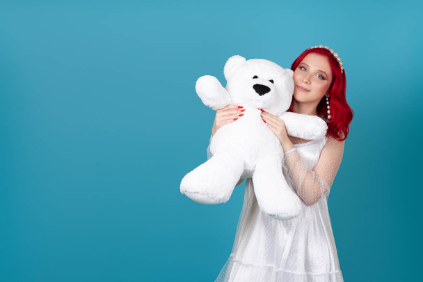 close-up a beautiful smiling woman in a white dress presses a large white teddy bear to her face , isolated on a blue background - Photo, Image