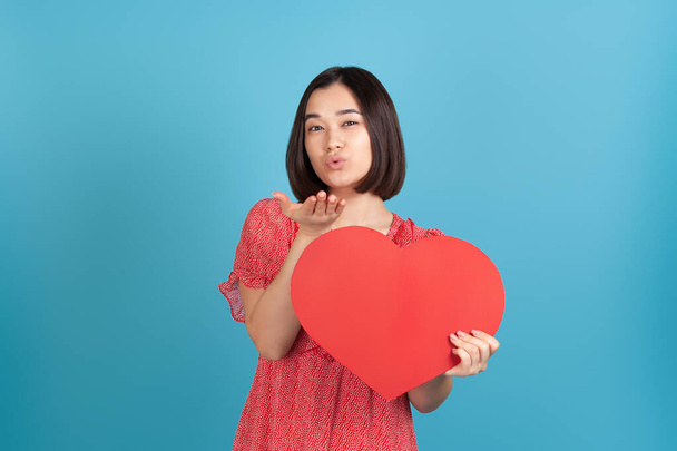 A romantic, lovesick young Asian woman in a red dress holds a large red paper heart and blows a kiss isolated on a blue background - Fotoğraf, Görsel
