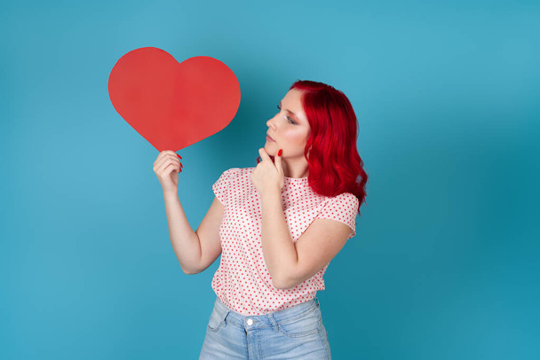 close-up pensive dreamy young woman with red hair holds a large red paper heart and looks at it isolated on a blue background - Foto, afbeelding