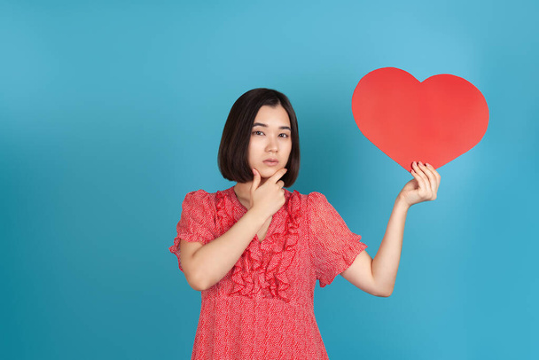 close-up thinking, decision-making young Asian woman in a red dress holds a large red paper heart in her hands and touches her chin with her hand , isolated on a blue background - Foto, Bild