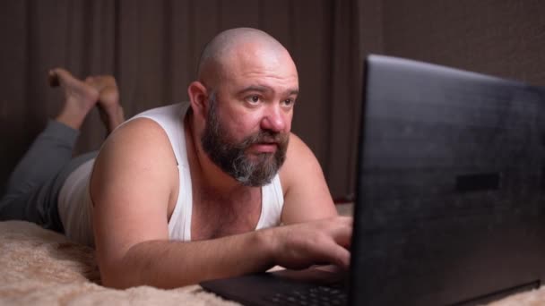 brodaty mężczyzna w T-shircie leży w domu na łóżku obok laptopa i intensywnie pisze coś na klawiaturze. Szczęśliwy człowiek odpoczywający w domu - Materiał filmowy, wideo