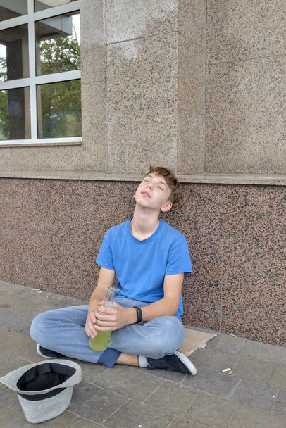 酔っぱらいのティーンエイジャーの乞食は彼の手に飲み物で座ってお金のために懇願します。十代のアルコール依存症の概念. - 写真・画像