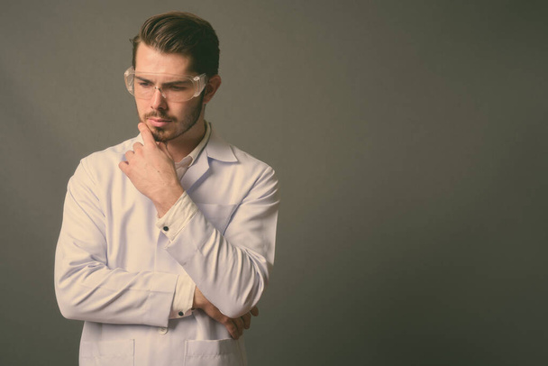 Giovane bello barbuto uomo medico indossa occhiali protettivi contro sfondo grigio - Foto, immagini