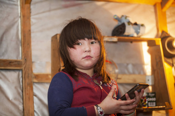 Ένα νεαρό κορίτσι, με τα εθνικά χειμωνιάτικα ρούχα των βόρειων κατοίκων της τούνδρας, βγάζει μια selfie σε ένα smartphone, το πρόβλημα της παιδικής τηλεφωνίας - Φωτογραφία, εικόνα