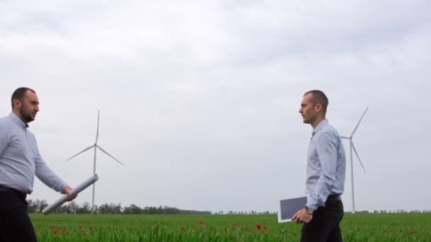 Deux hommes avec une tablette et un dessin en papier se serrent la main dans un champ vert sur fond de parcs éoliens en activité. Investissements dans l'énergie verte. - Séquence, vidéo