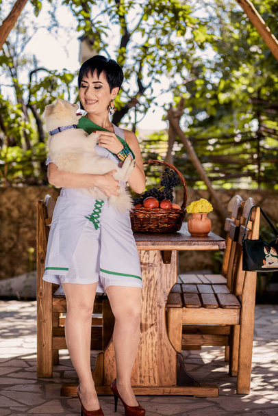 Красивая девушка обнимает своего белого щенка и стоит рядом со столом - Фото, изображение