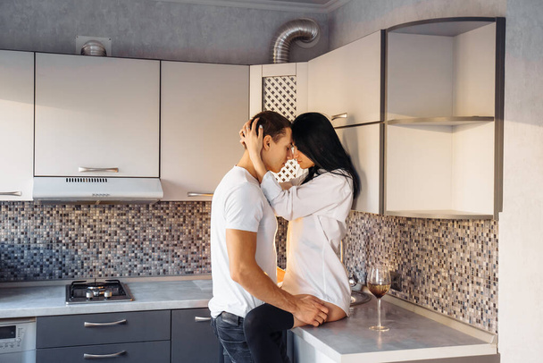 Cheerful active romantic couple of Caucasian ethnicity in underwear hug and kiss in kitchen. Happy carefree young europeans husband and wife enjoying weekend morning, laughing, chatting, having fun. - Photo, Image