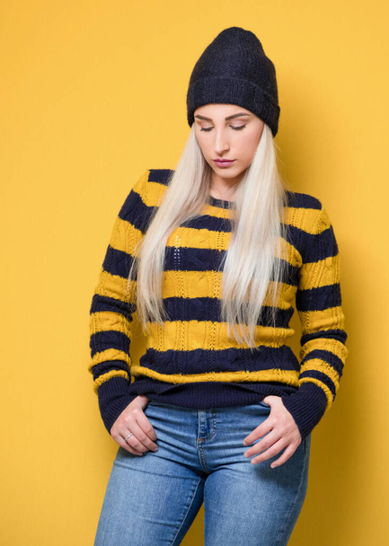 Young woman looking down and holding hands in pockets while standing and posing, model wearing woolen cap and sweater, isolated on yellow background. Full length of pretty girl. Studio shot - Φωτογραφία, εικόνα