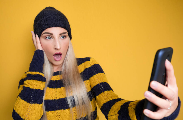 Scared young woman with mobile phone, model wearing woolen cap and sweater, isolated on yellow background. Shocked female person holding smartphone. Unbelievable. Studio shot - Foto, Imagen