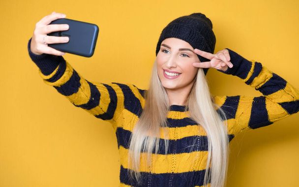 Selfie. Close up girl takes photo of himself with her smartphone, model wearing woolen cap and sweater, isolated on yellow background. Studio shot - Fotografie, Obrázek