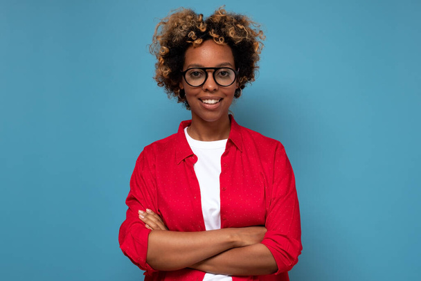 Χαμογελαστή νεαρή Αφρικανή με γυαλιά που στέκεται με σταυρωμένα τα χέρια στον μπλε τοίχο. - Φωτογραφία, εικόνα