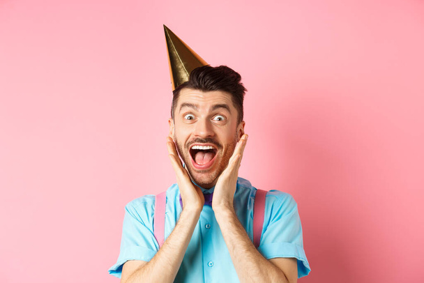 Holidays and celebration concept. Close-up of funny guy in birthday hat shouting surprised, screaming with joy, receiving awesome gift, standing happy on pink background - Photo, Image
