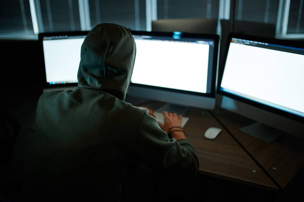 Male internet hacker in hood sitting at monitors, back view. Illegal web programmer at workplace, criminal occupation. Data hacking, cyber security - Foto, imagen