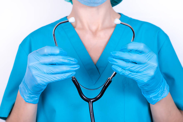 Female doctor in blue uniform and gloves holds a stethoscope in her hands. - Fotoğraf, Görsel