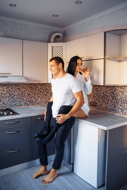 Attractive, hot, sexy brunette sits high on the kitchen furniture. Hugging boyfriend with legs passionately. Woman presses her body to the man. Dressed in white shirts and dark jeans. - Foto, Bild