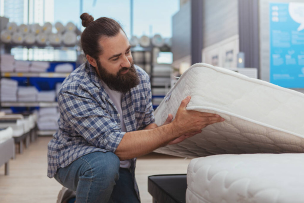Household shopping concept. Mature bearded man examining orthopedic mattress at furnishings store. Handsome male customer choosing new bed at furniture shop - Foto, afbeelding