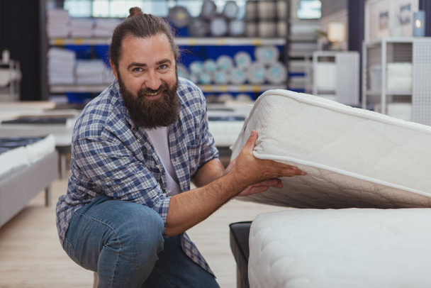 Healthy sleep concept. Handsome bearded mature man smiling to the camera, shopping for new orthopedic mattress at home goods store. Cheerful male customer choosing new bed - Fotografie, Obrázek