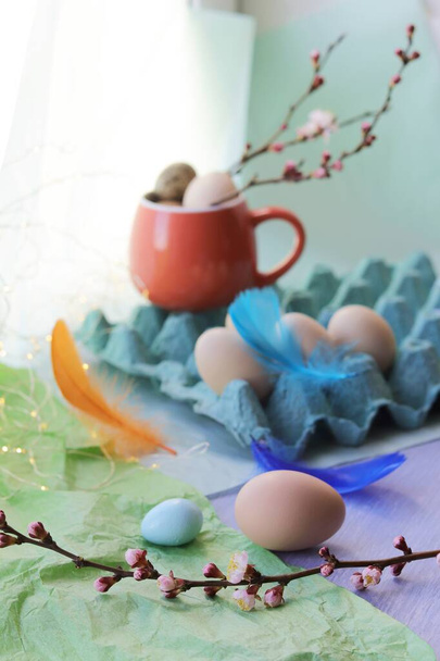 Los huevos de Pascua en el paquete y la taza, las plumas y las flores primaverales sobre la mesa sobre el fondo de la ventana  - Foto, imagen